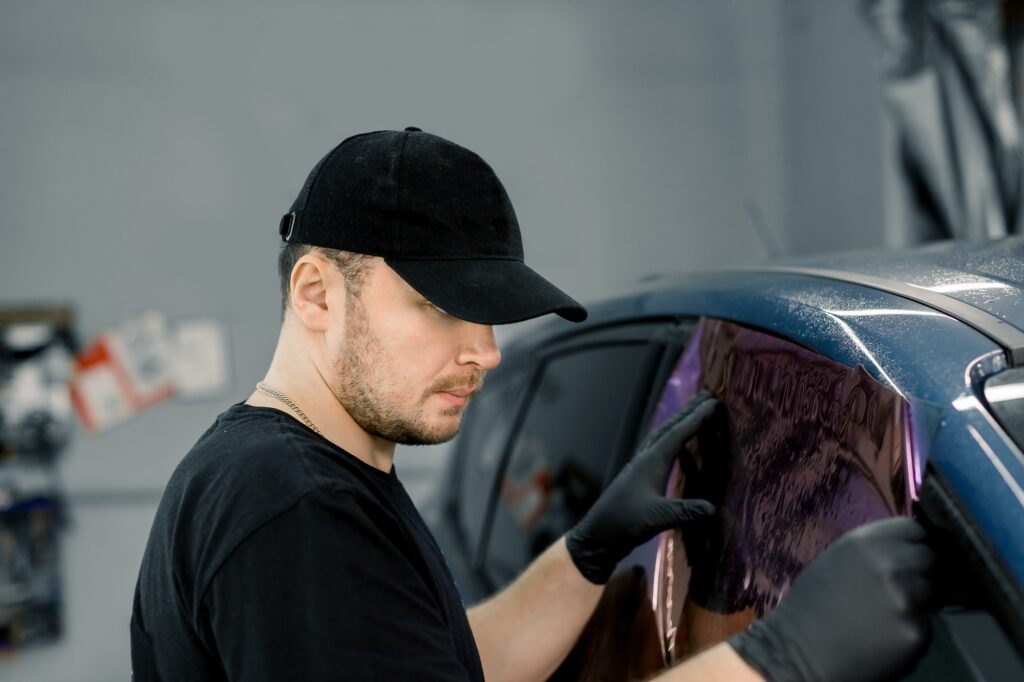 Professional car service worker wearing black cap and t-shirt, tinting a car window with tinted foil